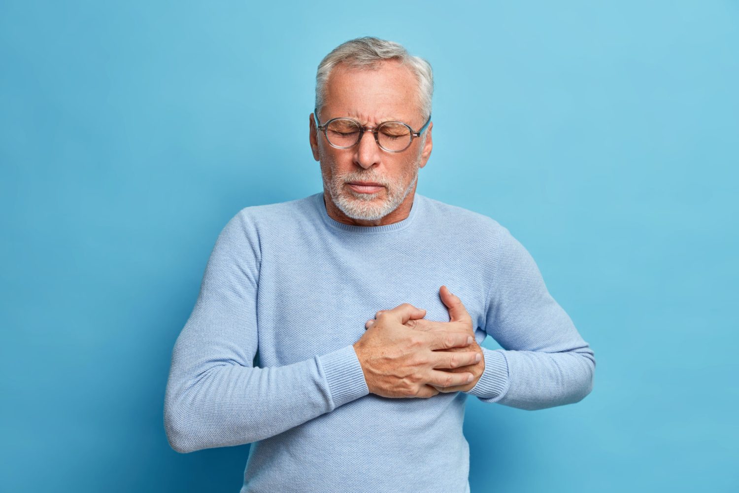 Senior man in spectacles presses hand to chest has heart attack suffers from unbearable pain closes eyes wears optical glasses poses against blue background. People age and problems with health